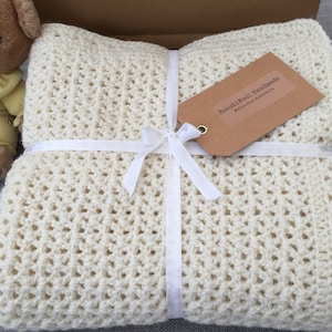 May include: A cream-colored crocheted blanket with a textured open-stitch pattern, folded and tied with a white satin ribbon. A brown rectangular paper tag with the text BeautiWool Handmade Melbourne Australia is attached to the ribbon.