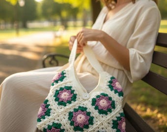 May include: A crochet tote bag constructed from off-white, dark green, and light purple granny squares featuring a floral motif. The bag has a single wide shoulder strap and a central white button closure at the top opening of the main compartment.