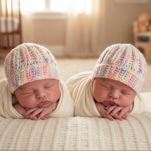 May include: Two newborn infants are sleeping side by side, each wearing a hand-knit beanie cap. The hats feature a ribbed texture and are crafted from variegated yarn in soft pastel shades of pink, yellow, blue, and white. Both infants are swaddled.