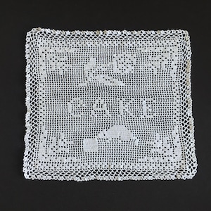 May include: White crocheted doily with a delicate floral pattern. The doily is square and has a scalloped edge.