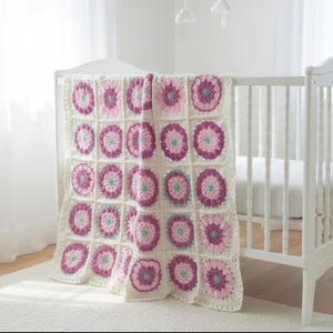 May include: A handmade crochet blanket featuring a repeating pattern of circular motifs in shades of magenta, light pink, and teal, joined together by an off-white yarn border. The blanket is draped over the side of a white wooden cot.