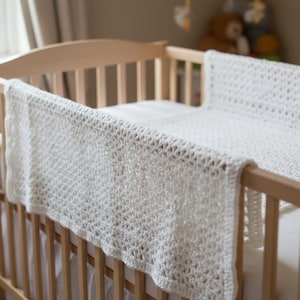 May include: A white crocheted baby blanket with a textured, open-weave pattern draped over the wooden rails of a crib. The blanket features a consistent, repeating stitch design throughout its rectangular shape, providing a soft, lightweight appearance.