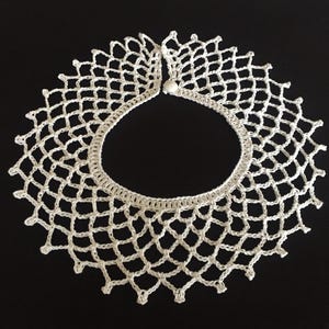 May include: A white crocheted collar featuring an intricate, open-work lattice pattern that radiates outward from a solid circular neckline. The piece is finished with a small, round white button closure at the top, creating a delicate, circular accessory.