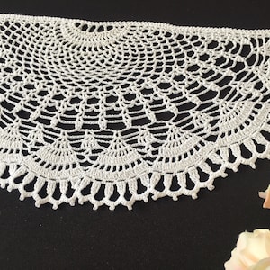 Puede incluir: Doily de ganchillo blanco con un diseño delicado e intrincado. El doily es circular con un borde festoneado.