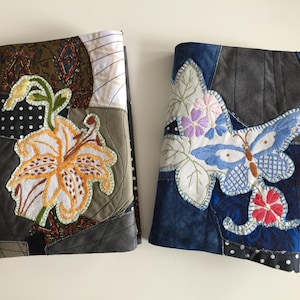 May include: Two rectangular fabric covers featuring intricate patchwork and embroidery. The left cover displays a large orange lily, while the right cover features a blue butterfly with floral accents. Both items are constructed from various quilted fabric pieces.