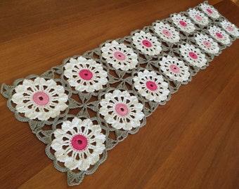 Könnte beinhalten: Ein rechteckiger Häkel-Tischläufer mit einem sich wiederholenden Blumenmuster. Jedes Motiv besteht aus einer leuchtend rosa Mitte, weißen Blütenblättern und einem hellgrauen Rand, der die einzelnen Blumenquadrate zu einem Stück verbindet.