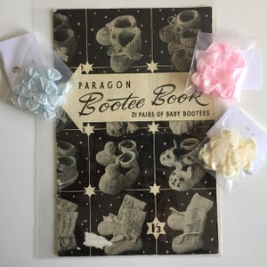 Puede incluir: Un libro vintage "Paragon Bootee Book" con ilustraciones de patucos de bebé. El libro está acompañado de pequeñas bolsas de lazos de satén azul claro, rosa y crema. La portada presenta un diseño en blanco y negro con estrellas.