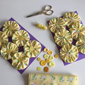 May include: A collection of yellow fabric yo-yos with a floral pattern arranged on two purple rectangular cards, accompanied by a pair of silver scissors, a spool of yellow thread, a variety of yellow buttons, and a folded piece of matching fabric.