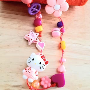 Peut inclure: Un collier de perles colorées avec divers breloques, dont un personnage Hello Kitty, des étoiles, des cœurs et des fleurs. Le collier est enfilé sur un cordon rouge et comprend des perles roses, jaunes et violettes. Le collier est sur une surface en bois.
