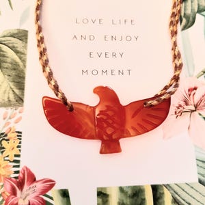 Könnte beinhalten: Eine rot-orangefarbene Adler-Anhänger-Halskette mit geflochtener Kordel. Der Anhänger ist mit detaillierten Flügeln und einem Kopf geschnitzt. Auf der Karte hinter der Halskette steht "LOVE LIFE AND ENJOY EVERY MOMENT". Der Hintergrund zeigt florale Muster.
