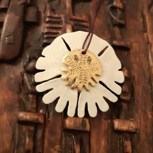 Pode incluir: Um pingente de cerâmica branca em forma de flor com um design de trilobite dourado no centro. O pingente está pendurado em um cordão marrom.