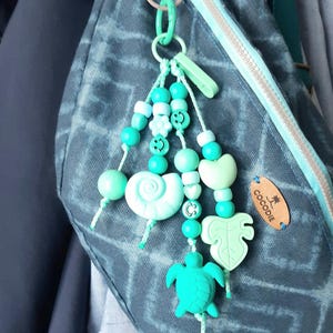 Könnte beinhalten: Ein türkis-weißes Perlen-Accessoire mit einem Schildkröten-, Blatt- und Muscheldesign. Der Anhänger ist an einer Tasche mit einem "COCODIE"-Etikett befestigt. Der Anhänger enthält Smiley-Perlen und einen grünen Verschluss.