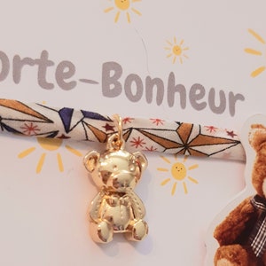 Peut inclure: Un bracelet en tissu avec un motif géométrique et un charme en forme d'ours en or. Le bracelet est étiqueté "Porte-Bonheur", qui signifie "porte-bonheur" en français.