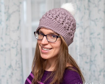 May include: A woman wearing a purple crocheted hat with a white pom pom on top. She is wearing glasses and a purple and white striped shirt. The text "nicole chase" is visible in the bottom right corner of the image.