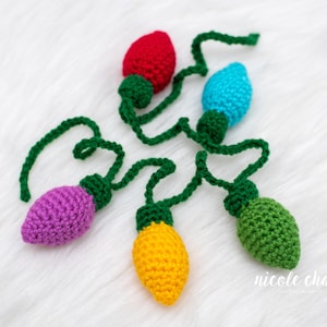 May include: A string of five crocheted Christmas lights in red, blue, purple, yellow, and green. Each light has a green base and is connected by a green crocheted wire. The lights are arranged on a white, fluffy surface.