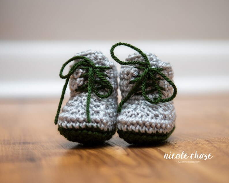May include: A pair of gray crocheted baby booties with green laces. The booties have a green trim around the bottom.