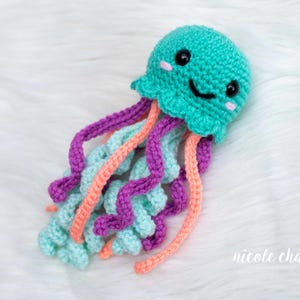 May include: A crocheted jellyfish toy in shades of teal, purple, and coral. The jellyfish has a smiling face with black eyes and pink cheeks. The tentacles are a mix of colors and hang down. The toy is handmade and sits on a white, fluffy surface.