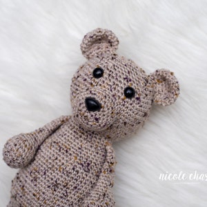May include: A crocheted teddy bear with brown and purple yarn, black button eyes, and a black nose. The bear is lying on a white fluffy surface.