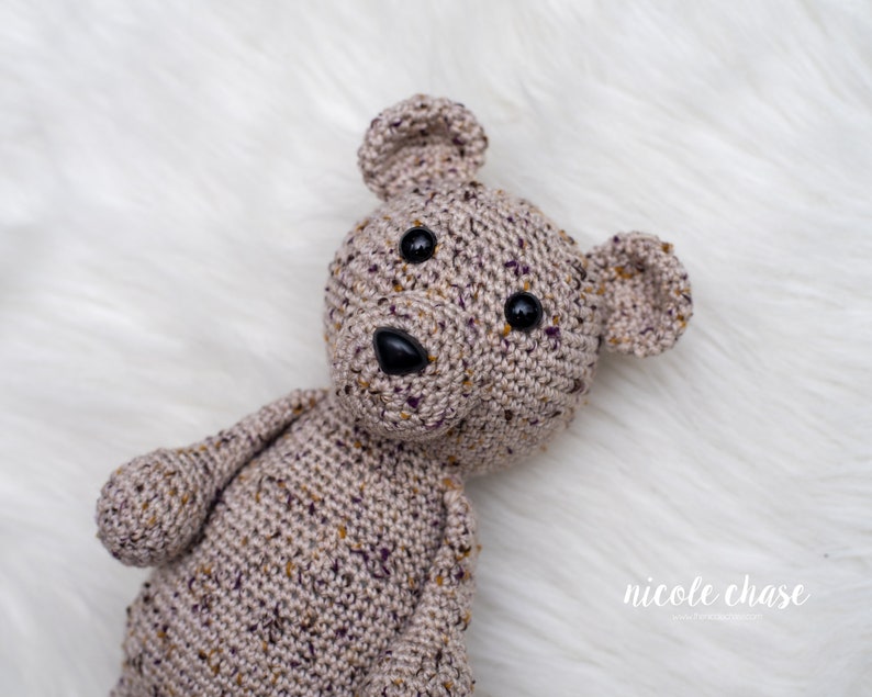 May include: A crocheted teddy bear with brown and purple yarn, black button eyes, and a black nose. The bear is lying on a white fluffy surface.