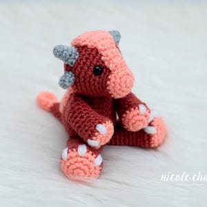 May include: A handmade crochet dragon in shades of red, coral, and gray. The plush toy features a detailed design with horns, paws, and a tail. The dragon is sitting on a white, fluffy surface.