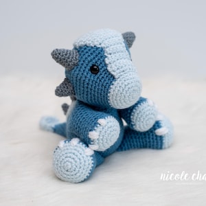Può includere: Un drago all'uncinetto fatto a mano nei toni del blu e del grigio. Il peluche ha la testa e la pancia azzurro chiaro, con accenti blu scuro e grigi sulle corna, sulla schiena e sui piedi. Il drago è seduto su una superficie bianca e soffice.