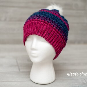 May include: A crocheted beanie hat with a pom pom on top. The hat is striped in shades of pink, purple, and blue.