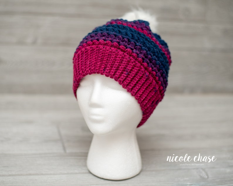 May include: A crocheted beanie hat with a pom pom on top. The hat is striped in shades of pink, purple, and blue.