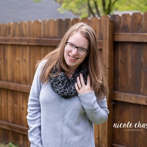 K&ouml;nnte beinhalten: Eine Frau in einem grauen Pullover und einem schwarz-wei&szlig;en Infinity-Schal l&auml;chelt in die Kamera. Sie tr&auml;gt eine Brille und hat lange braune Haare.