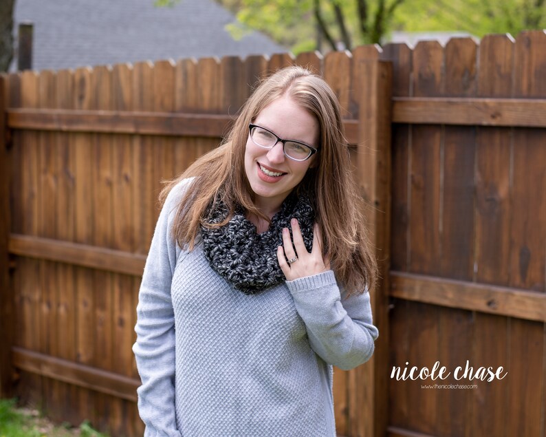 K&ouml;nnte beinhalten: Eine Frau in einem grauen Pullover und einem schwarz-wei&szlig;en Infinity-Schal l&auml;chelt in die Kamera. Sie tr&auml;gt eine Brille und hat lange braune Haare.