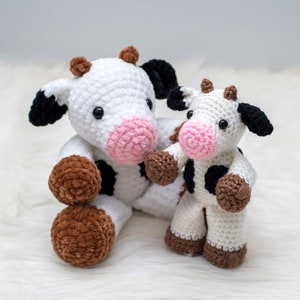 Op de afbeelding: Twee gehaakte knuffelkoeien, een groter dan de andere. De grotere koe is wit met zwarte vlekken en bruine hoeven. De kleinere koe is wit met zwarte vlekken en bruine hoeven. Beide koeien hebben een roze neus en zwarte ogen.