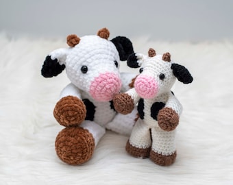 Puede incluir: Dos peluches de vaca de crochet, uno más grande que el otro. La vaca más grande es blanca con manchas negras y pezuñas marrones. La vaca más pequeña es blanca con manchas negras y pezuñas marrones. Ambas vacas tienen la nariz rosa y los ojos negros.