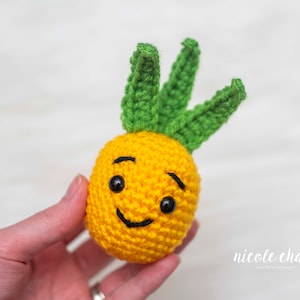 Könnte beinhalten: Ein gehäkeltes Ananas-Spielzeug mit einem lächelnden Gesicht, grünen Blättern und schwarzen Augen.