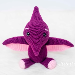 May include: A purple crocheted pterodactyl toy with pink wings and black eyes. The toy is sitting on a white surface.