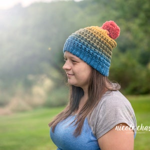 May include: A woman wearing a colorful crocheted beanie with a red pompom. The beanie has shades of blue, green, yellow, and orange. The woman is wearing a blue and gray shirt and has long brown hair.