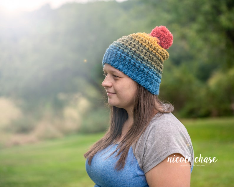 May include: A woman wearing a colorful crocheted beanie with a red pompom. The beanie has shades of blue, green, yellow, and orange. The woman is wearing a blue and gray shirt and has long brown hair.