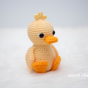 May include: A crocheted yellow duck with orange feet and black eyes. The duck is sitting on a white fluffy surface.