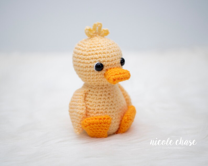 May include: A crocheted yellow duck with orange feet and black eyes. The duck is sitting on a white fluffy surface.
