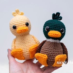 May include: Two crocheted stuffed toys, a yellow duck and a brown duck with a green head, sitting in a person's hand.