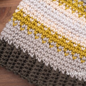 Pode incluir: Close-up de um chap&eacute;u de croch&ecirc; com listras de fio bege, creme e amarelo mostarda. O chap&eacute;u tem uma borda de croch&ecirc; marrom escura.