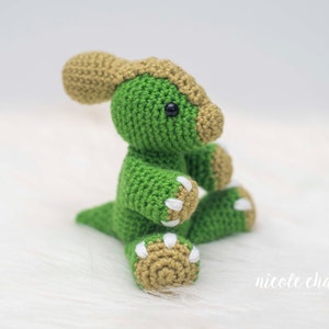 May include: A handmade green and tan crochet dinosaur toy. The dinosaur has a green body, tan head crest, and tan feet with white claws. The toy is sitting on a white, fluffy surface. The dinosaur has black safety eyes.
