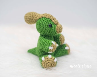 May include: A handmade green and tan crochet dinosaur toy. The dinosaur has a green body, tan head crest, and tan feet with white claws. The toy is sitting on a white, fluffy surface. The dinosaur has black safety eyes.