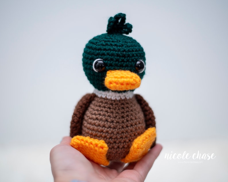 May include: A crocheted stuffed duck toy with green, brown, white, and orange yarn. The duck has black eyes and an orange beak.