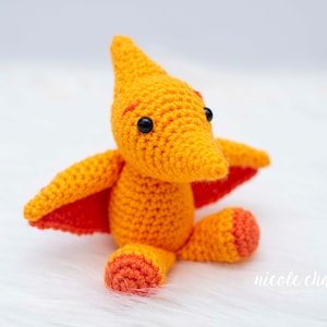 May include: A crocheted orange and red pterodactyl toy with black eyes. The toy is sitting on a white fluffy surface.