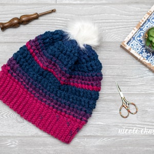 May include: A crocheted beanie hat with a pink and blue ombre pattern and a white pom pom on top. The hat is lying on a gray wooden surface.