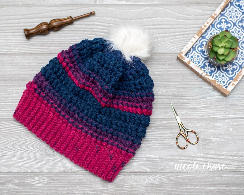May include: A crocheted beanie hat with a pink and blue ombre pattern and a white pom pom on top. The hat is lying on a gray wooden surface.