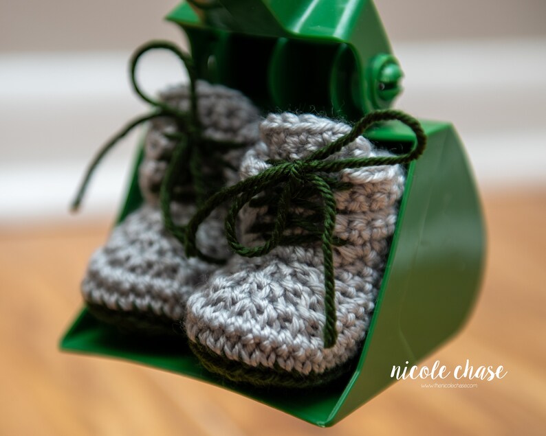 May include: A pair of gray crocheted baby booties with green laces. The booties are sitting in a green plastic scoop.