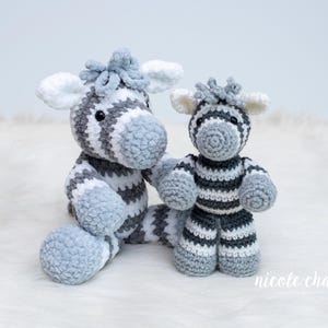 May include: Two handmade crochet zebra soft toys. The larger zebra is seated, with grey and white stripes, and a blue-grey mane and feet. The smaller zebra is standing, with similar colouring. The background is a soft white.