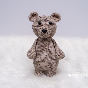 May include: A crocheted brown and beige teddy bear with black eyes. The bear is standing on a white fluffy surface.