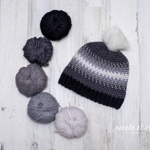 Pode incluir: Um gorro de croch&ecirc; em degrad&ecirc; cinza e branco com um pompom branco. O gorro &eacute; cercado por novelos de l&atilde; em v&aacute;rios tons de cinza.