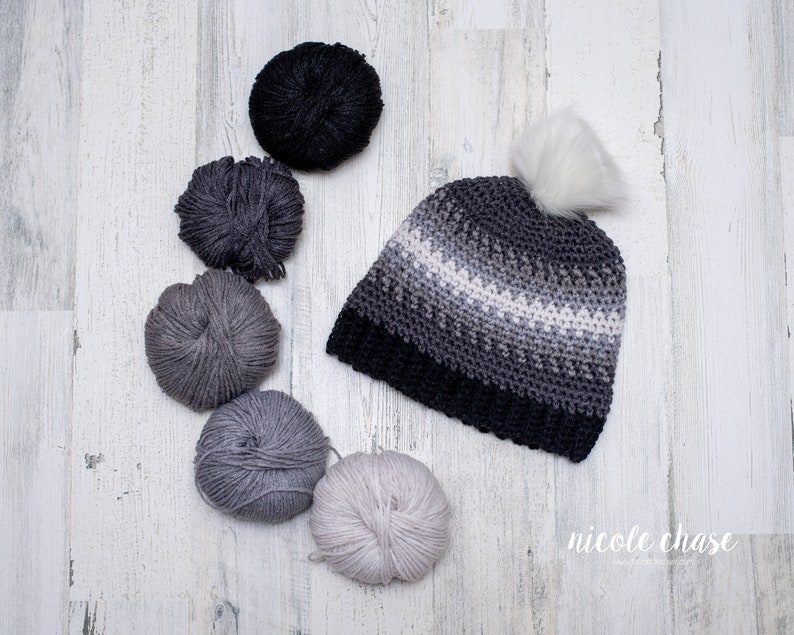 Pode incluir: Um gorro de croch&ecirc; em degrad&ecirc; cinza e branco com um pompom branco. O gorro &eacute; cercado por novelos de l&atilde; em v&aacute;rios tons de cinza.